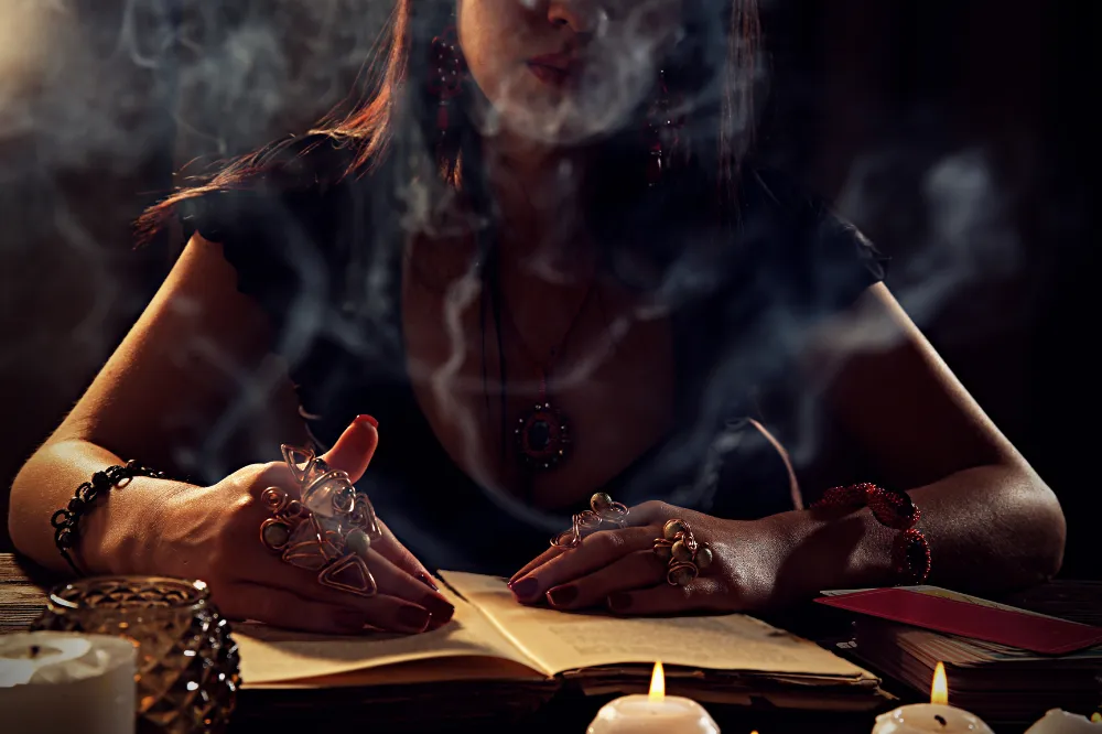 A person with long hair is seated at a table, writing in an open notebook surrounded by candles, with smoke swirling in the air, creating a mystical atmosphere. The setting suggests a spiritual or psychic reading session.