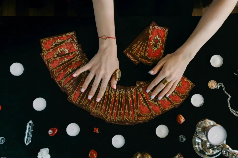 A pair of hands gently spreads a deck of tarot cards on a dark surface, surrounded by crystals and candles, suggesting a spiritual or psychic reading session. The atmosphere is calm and mystical.