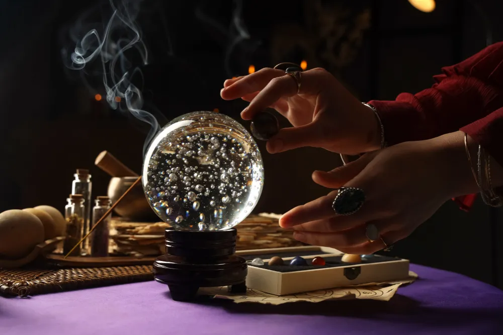 A pair of hands hovers over a crystal ball surrounded by smoke, set on a purple cloth with various mystical tools nearby. The scene evokes a sense of mystery and spirituality.